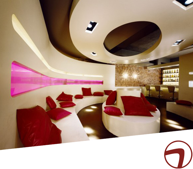 Modernes Lounge-Interieur im Vollblut OneNinetyEast mit geschwungenen weißen Sitzgelegenheiten, roten Kissen, rosa beleuchteter Wanddekoration, kreisförmiger Deckengestaltung und einem stilvollen Barbereich mit Flaschen vor einer unverputzten Ziegelwand.